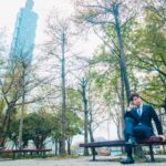 台北101と緑豊かな公園でベンチに座り電話する紺色スーツの男性
